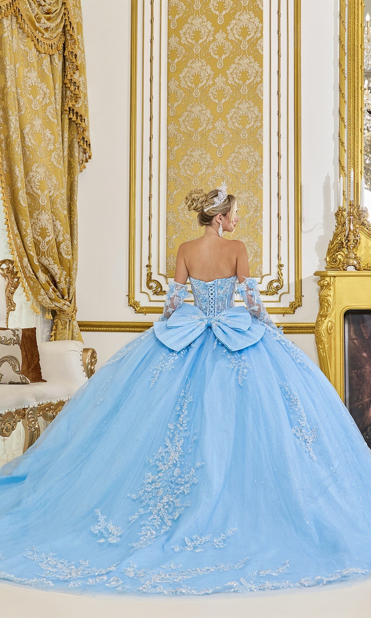A woman in the Quinceanera Dress 5081 by Dancing Queenโfeaturing glitter tulle, beaded lace appliquรฉ, and a large bowโstands in an ornate gold-accented room, facing away from the camera.